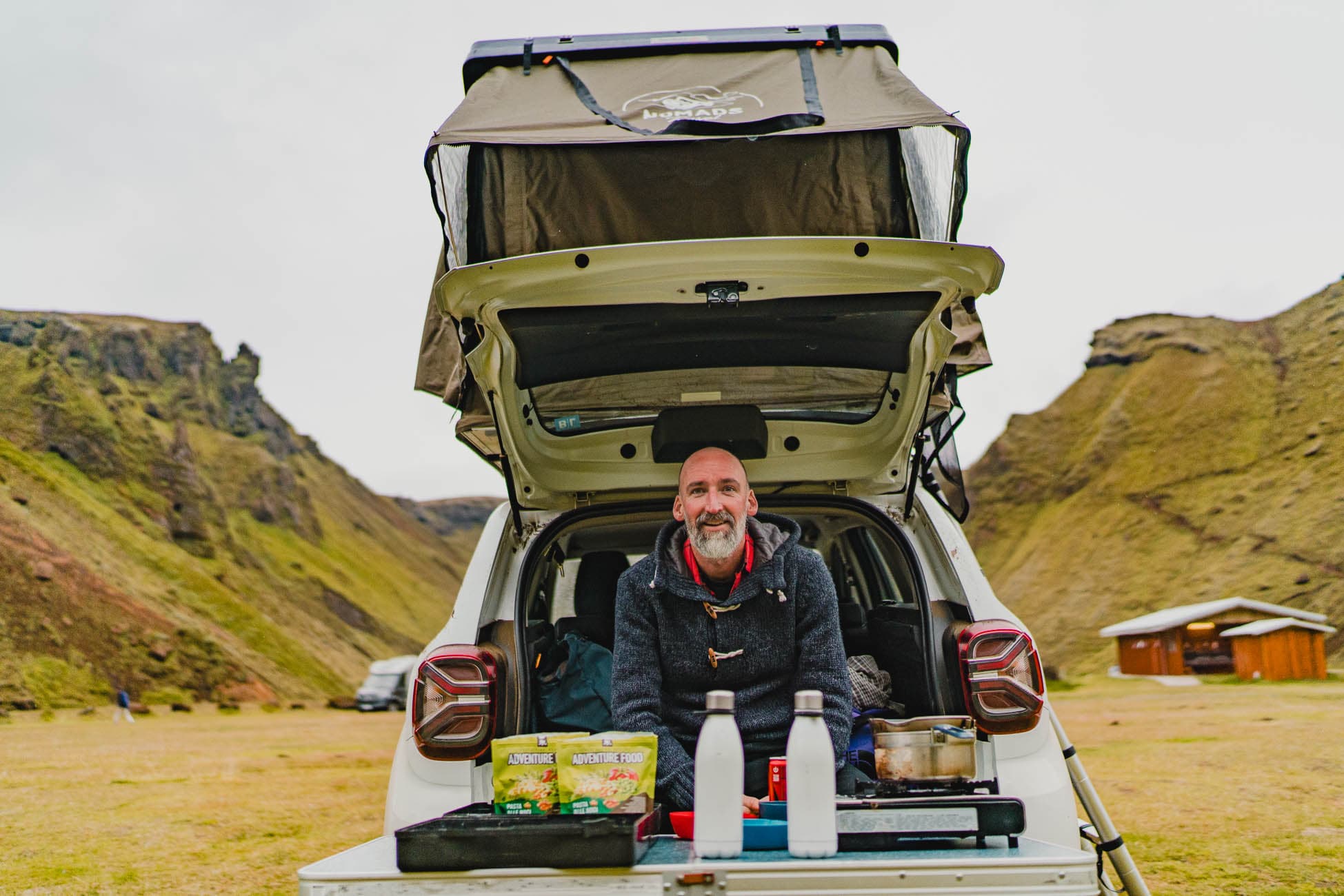 Een man zit achterin een auto met open kofferbak, klaar om te kamperen in IJsland, met kookgerei en eten op een tafel. De auto heeft een daktent, genesteld in een met gras begroeide vallei die de groei van de natuur laat zien, met heuvels en een hut in de buurt.