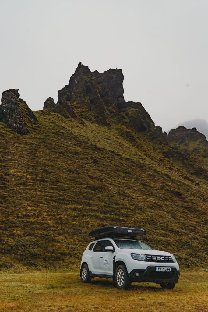 Een witte SUV met dakkoffer staat geparkeerd op een met gras begroeid terrein aan de voet van een steile, rotsachtige heuvel in IJsland, onder een bewolkte lucht die de wilde schoonheid en natuurlijke groei van het landschap vastlegt.