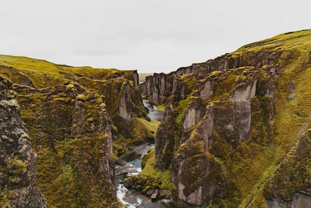 Een diepe, smalle canyon met steile, met mos bedekte kliffen en een kronkelende rivier die door het midden stroomt, onder een bewolkte IJsland-hemel - een opvallend landschap waar de groei van de natuur volop te zien is.