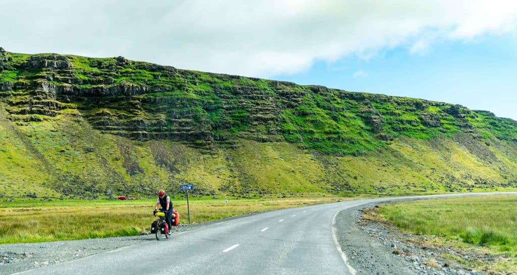Een fietser met volle fietstassen staat bij een verkeersbord op een kronkelige landweg in IJsland. Op de achtergrond rijzen steile groene kliffen op tegen een gedeeltelijk bewolkte lucht, die het open, grasachtige landschap en de natuurlijke groei van de regio laat zien.