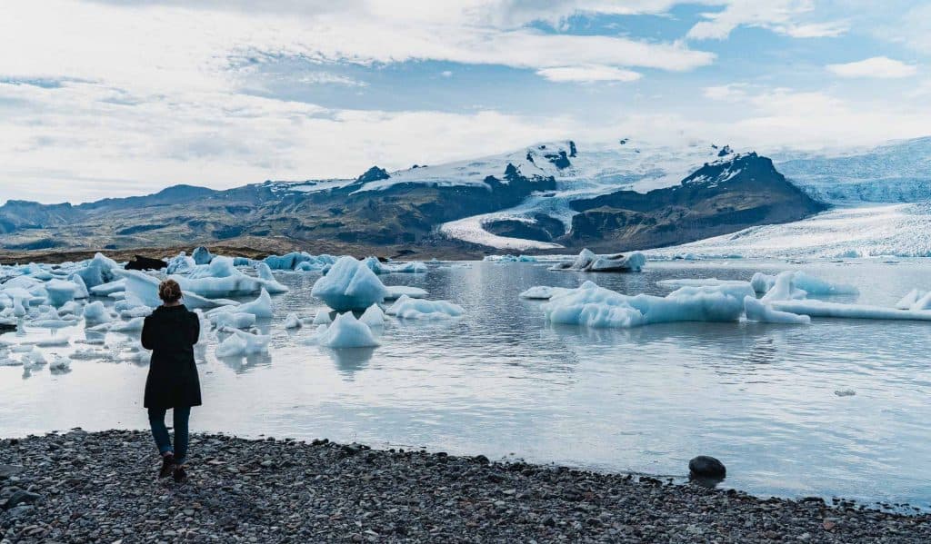 Een persoon in een donkere jas staat op een rotsachtige kust in IJsland en kijkt uit op drijvende ijsbergen in een gletsjerlagune, omringd door bergen met besneeuwde toppen en een bewolkte hemel - een scène die de rauwe schoonheid en groei van de natuur weerspiegelt.