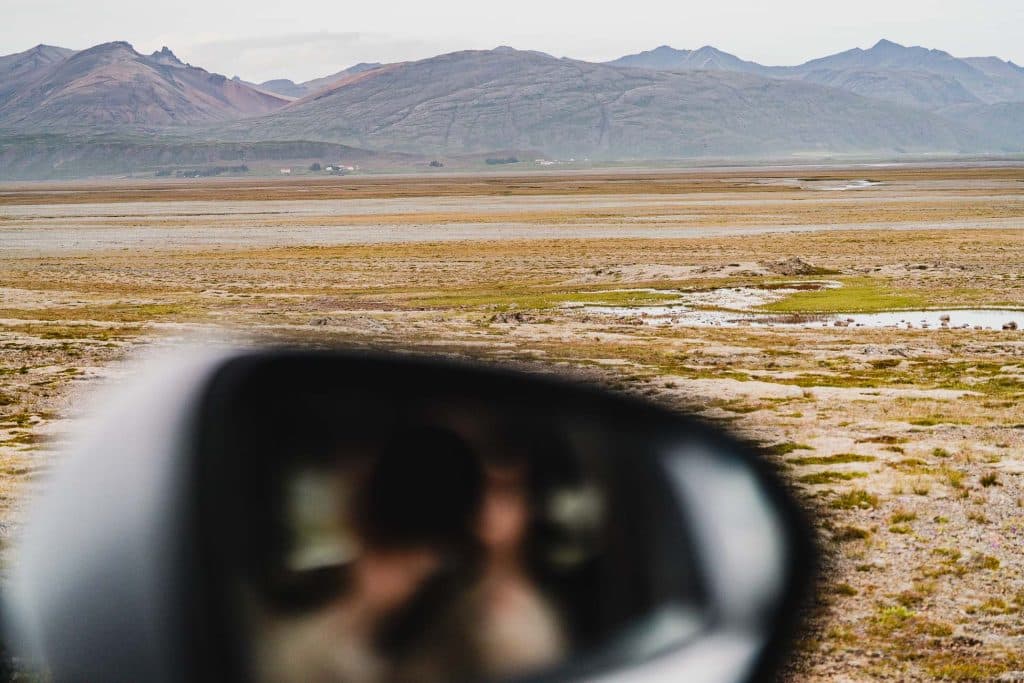Een droge grasvlakte met bergen op de achtergrond onder een bewolkte hemel. De vage weerspiegeling van een persoon is zichtbaar in de zijspiegel van een auto, wat de geest van IJslandse nomaden oproept terwijl ze naar het weidse landschap staren.