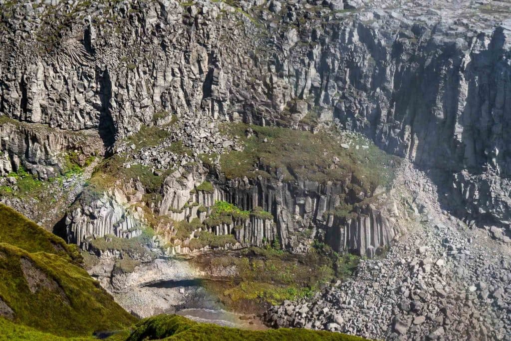 Steile rotsachtige kliffen met zuilen van basaltformaties in IJsland, vlekken groen mos die groei symboliseren en verspreide rotsen aan de voet; linksonder verschijnt een vage regenboog.