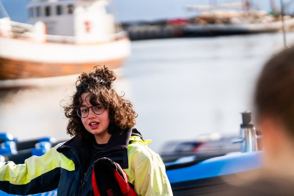 Een jong persoon met krullend haar en een bril, die een geel en zwart jack draagt, staat bij het water met boten op de achtergrond op een zonnige dag in IJsland, een moment van groei en avontuur vastleggend.
