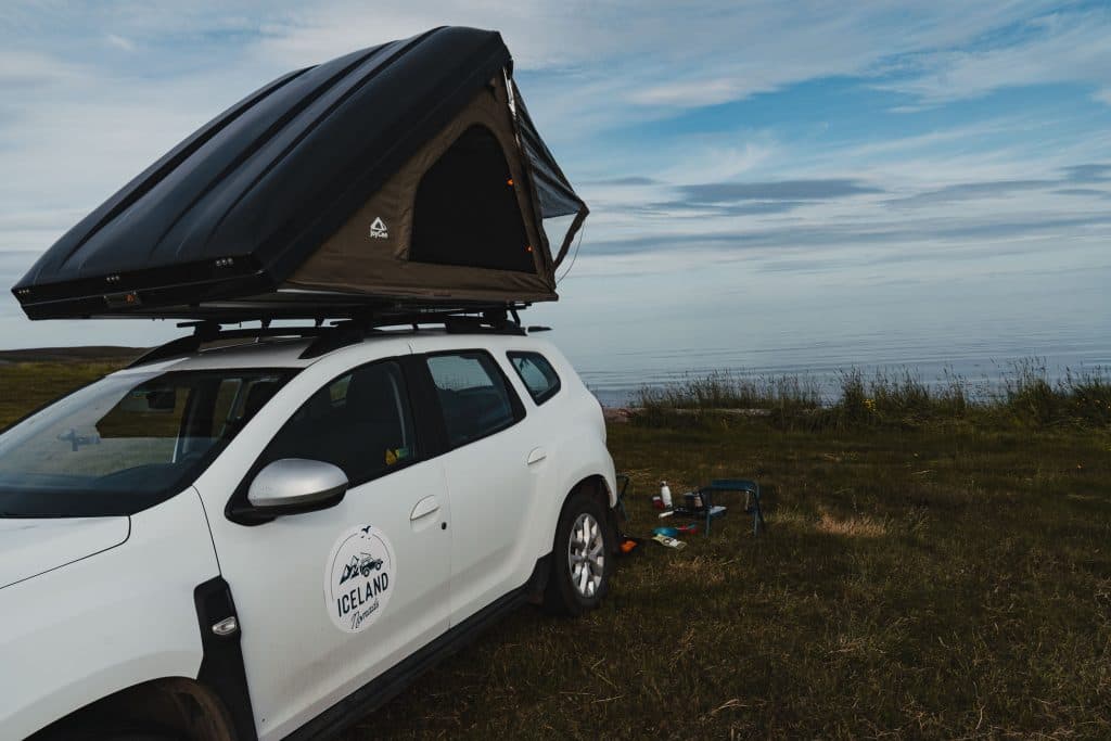 Een witte SUV met daktent staat geparkeerd op een grasveld bij de kalme zee in IJsland. Kampeerspullen, waaronder een fornuis en waterkoker, staan naast de auto. De lucht is gedeeltelijk bewolkt met blauwe tinten en de avontuurlijke geest groeit.