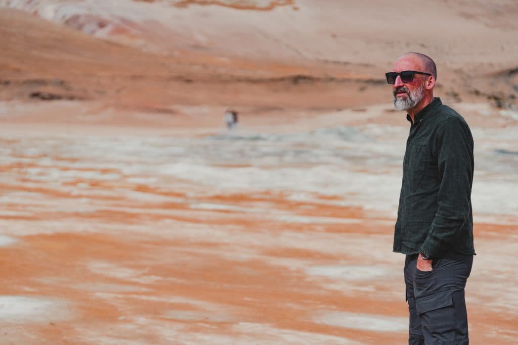 Een man met een zonnebril en donkere kleding staat met zijn handen in zijn zakken op een dor, oranje en wit IJsland-landschap, dat groei symboliseert tegen de grimmige heuvels op de achtergrond.