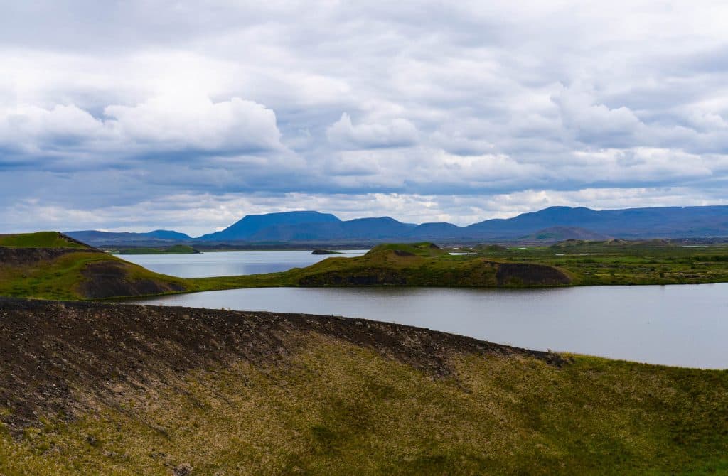 Een schilderachtig uitzicht op een meer in IJsland omringd door groene heuvels onder een bewolkte hemel, met bergen in de verte op de achtergrond.