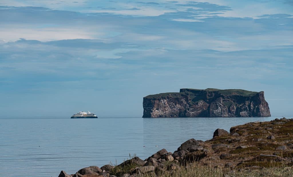 Een veerboot vaart over kalm blauw water nabij een groot, rotsachtig eiland met grastoppen in IJsland, gezien vanaf een rotsachtige kustlijn onder een gedeeltelijk bewolkte hemel, en legt de serene schoonheid en natuurlijke groei van het landschap vast.