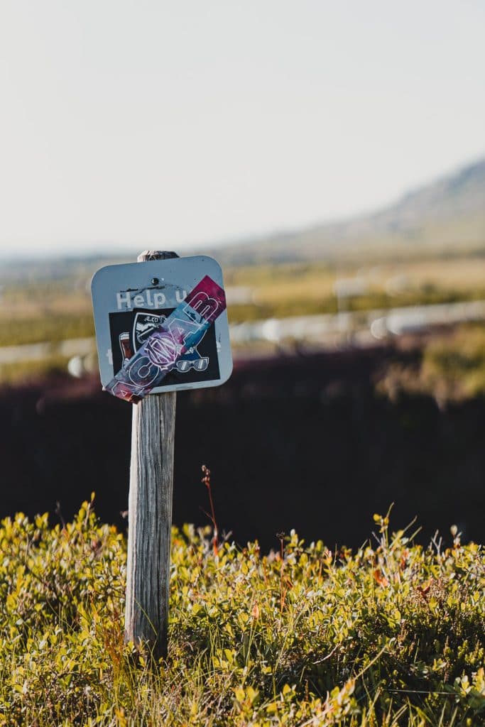Een verweerd "Help ons"-bord op een houten paal staat in een grasveld, gedeeltelijk bedekt door een snoeppapiertje. Op de achtergrond wijst een vage weg op een avontuurlijke Roadtrip IJsland onder een heldere hemel.