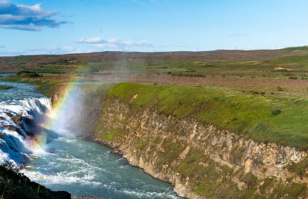 Een rivier stroomt door een rotsachtige kloof met groen gras bovenop, terwijl een regenboog glinstert in de mist - een onvergetelijk gezicht tijdens een 7-daagse roadtrip met Iceland Nomads onder de helderblauwe hemel.