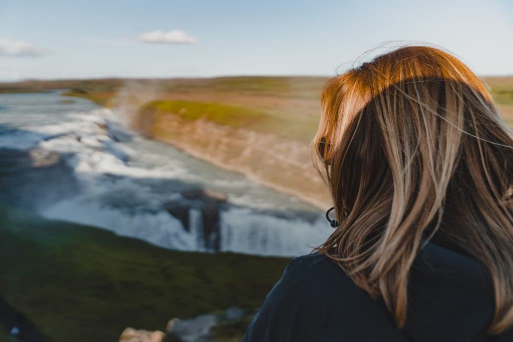 Een persoon met lichtbruin haar staat uit te kijken over een brede, krachtige waterval die neerklettert over een groen landschap - perfect decor voor een 7-daagse roadtrip met Iceland Nomads, mist die oprijst onder de blauwe hemel.