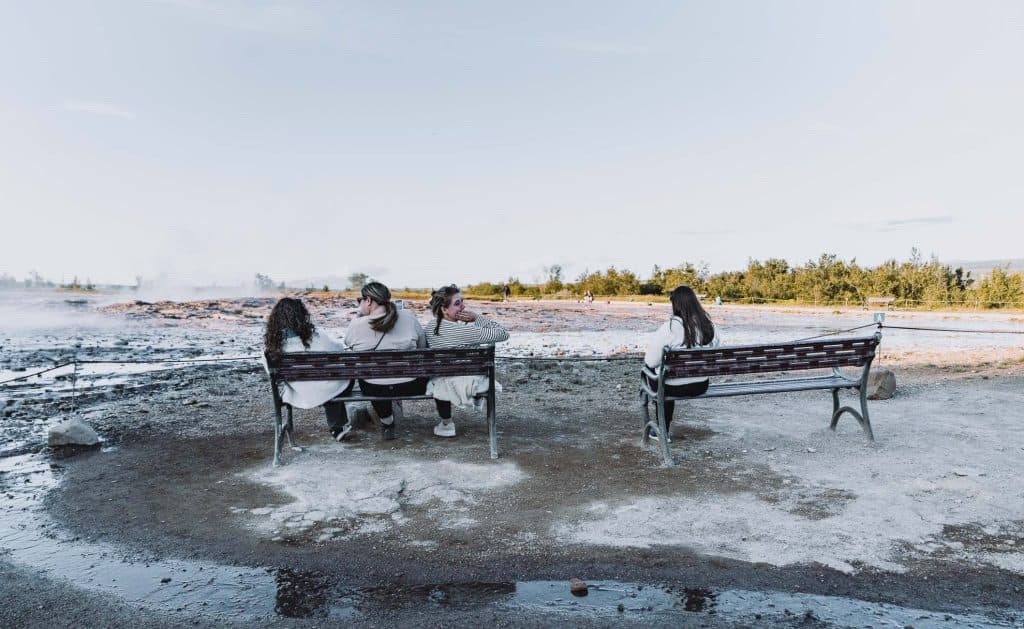 Vier mensen, die deel uitmaken van de Iceland Nomads op een 7-daagse roadtrip, zitten op bankjes die uitkijken op een prachtig, rotsachtig landschap met water en bomen - drie samen op één bankje en één alleen - en genieten allemaal van het uitzicht van Roadtrip IJsland.