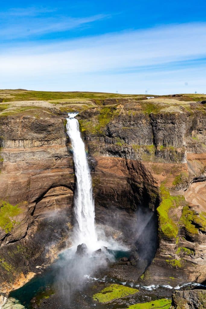 Een hoge waterval stort zich over een rotsachtige klif in een mistige poel eronder, omgeven door groen gras en ruig bruin terrein - een adembenemende stop tijdens uw 7-daagse roadtrip met Iceland Nomads onder een helderblauwe hemel met lichte wolken.