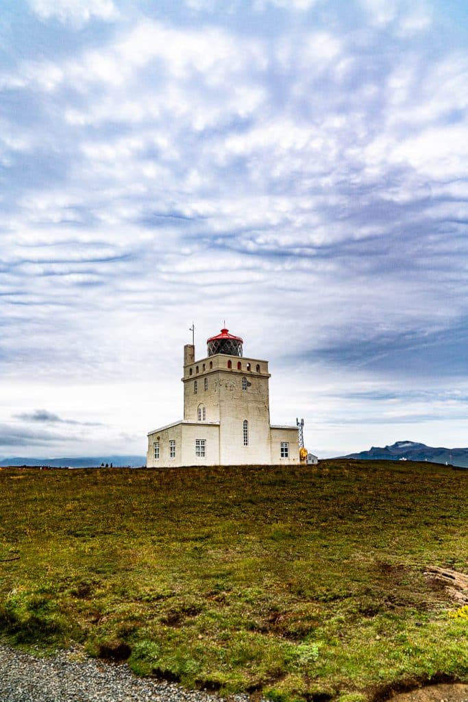 Een witte vuurtoren met een rode koepel staat op open groen gras onder een bewolkte hemel, met bergen in de verte op de achtergrond - een perfecte stop tijdens uw 7-daagse roadtrip met Iceland Nomads.
