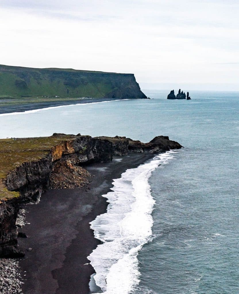 Rotsachtige kliffen en een zwart zandstrand ontmoeten de oceaan, met witte golven die langs de kust slaan; verre zeebergen rijzen op uit het water onder een bewolkte hemel - een perfect tafereel voor uw 7-daagse Roadtrip IJsland met Iceland Nomads.