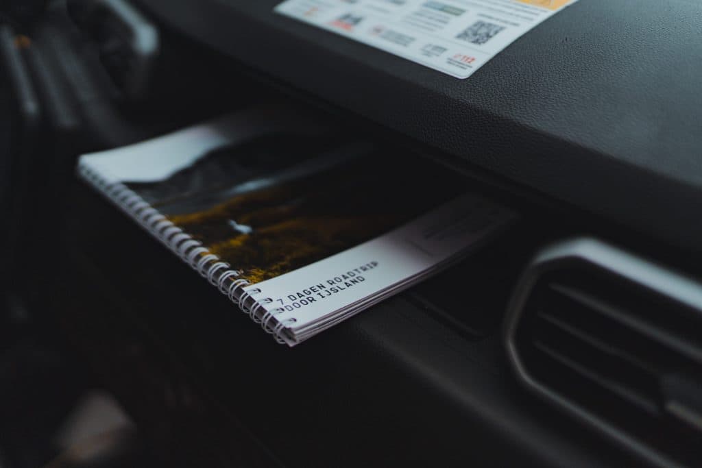Een notitieboekje met spiraalbodem dat gedeeltelijk in het dashboardkastje van een auto is verstopt, met een omslag met een landschapsafbeelding en de tekst "3 DAGEN ROADTRIP DOOR IJSLAND," perfect om je eigen Roadtrip IJsland te plannen of om je aan te sluiten bij de Iceland Nomads.