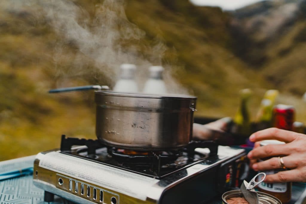 Een metalen pan met opstijgende stoom staat op een draagbaar gasfornuis buiten tijdens een 7-daagse roadtrip, met een hand die een blikje opent vlakbij. De achtergrond toont een onscherp natuurlijk landschap met groen en heuvels.