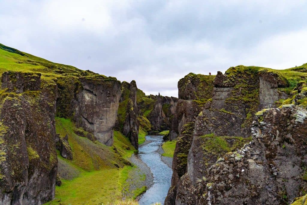 Een smalle rivier kronkelt door een dramatische kloof met steile, met mos bedekte kliffen onder een bewolkte hemel - een weelderig en ruig landschap dat perfect is voor een onvergetelijke 7-daagse roadtrip met Iceland Nomads.