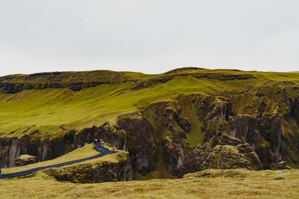 Mensen lopen over een smal pad langs een met gras begroeide klif met steile rotsformaties en bemoste heuvels onder een bewolkte hemel, waarbij de wilde schoonheid perfect is voor een IJsland Nomads avontuur of een 7-daagse roadtrip.