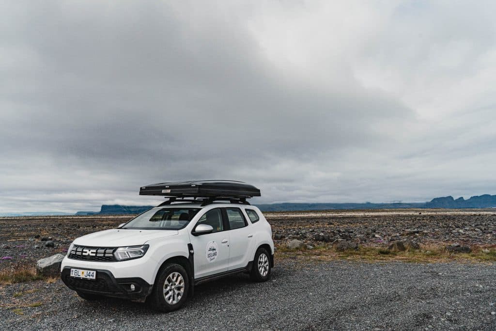 Een witte SUV met dakkoffer staat geparkeerd op grind te midden van het ruige landschap van IJsland, perfect voor een 7-daagse roadtrip met Iceland Nomads. Bergen doemen op in de verte onder een bewolkte hemel, die de geest van een roadtrip IJsland vastleggen.