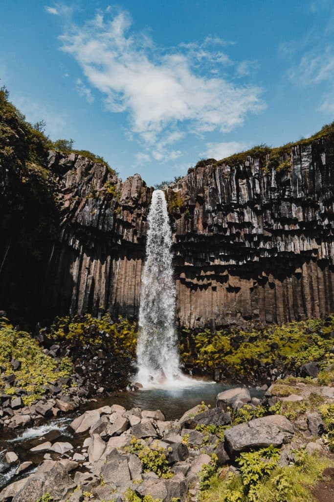 Een hoge waterval stort zich over dramatische zwarte basaltzuilen in een rotsachtige poel, omgeven door groen mos. Beleef dit adembenemende schouwspel onder een blauwe hemel met Iceland Nomads tijdens een 7-daagse roadtrip door de natuurwonderen van IJsland.