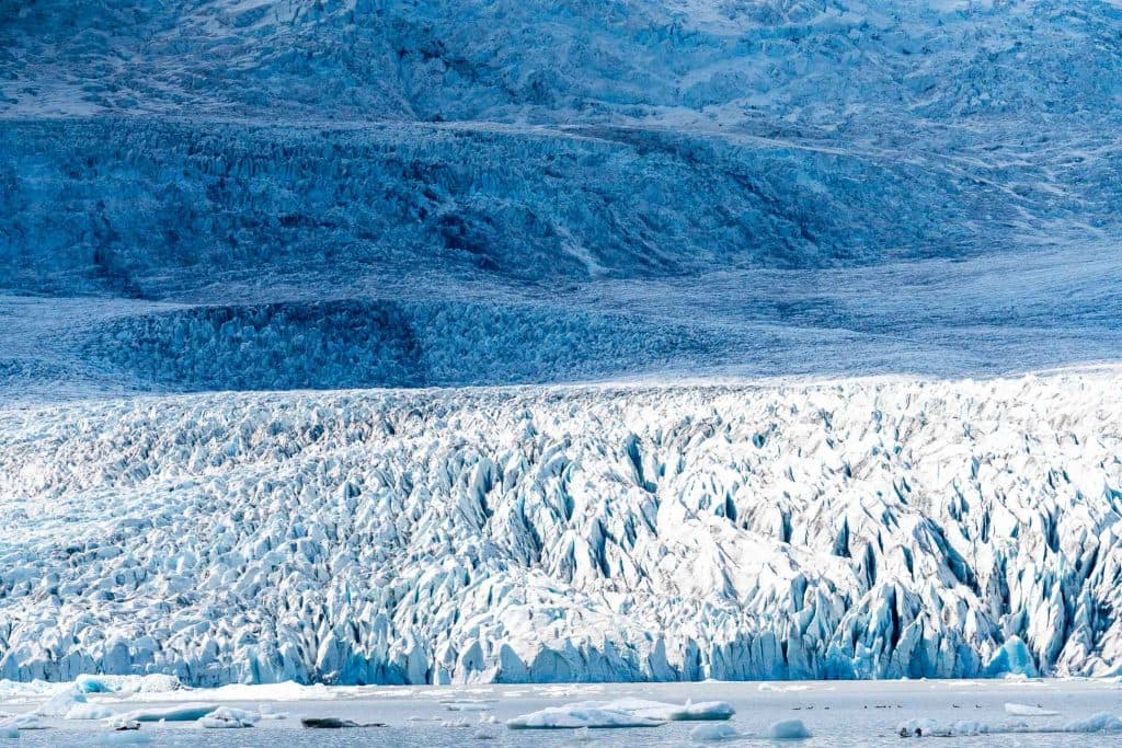 Een gletsjer met grillige ijsformaties aan de voorkant en gelaagde besneeuwde oppervlakken op de achtergrond, allemaal in blauwe en witte tinten - een iconisch gezicht op elke Roadtrip IJsland of avontuur met Iceland Nomads - plus drijvende stukken ijs op het water aan de basis.