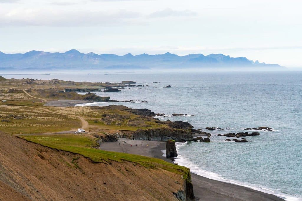 Uitzicht op een rotsachtige kustlijn met een zwart zandstrand, met gras begroeide kliffen en bergen in de verte onder een bewolkte hemel - perfect voor een 7-daagse Roadtrip IJsland. Een paar auto's en mensen zijn te zien in de buurt van de kust, die de geest van IJsland Nomaden vastlegt.