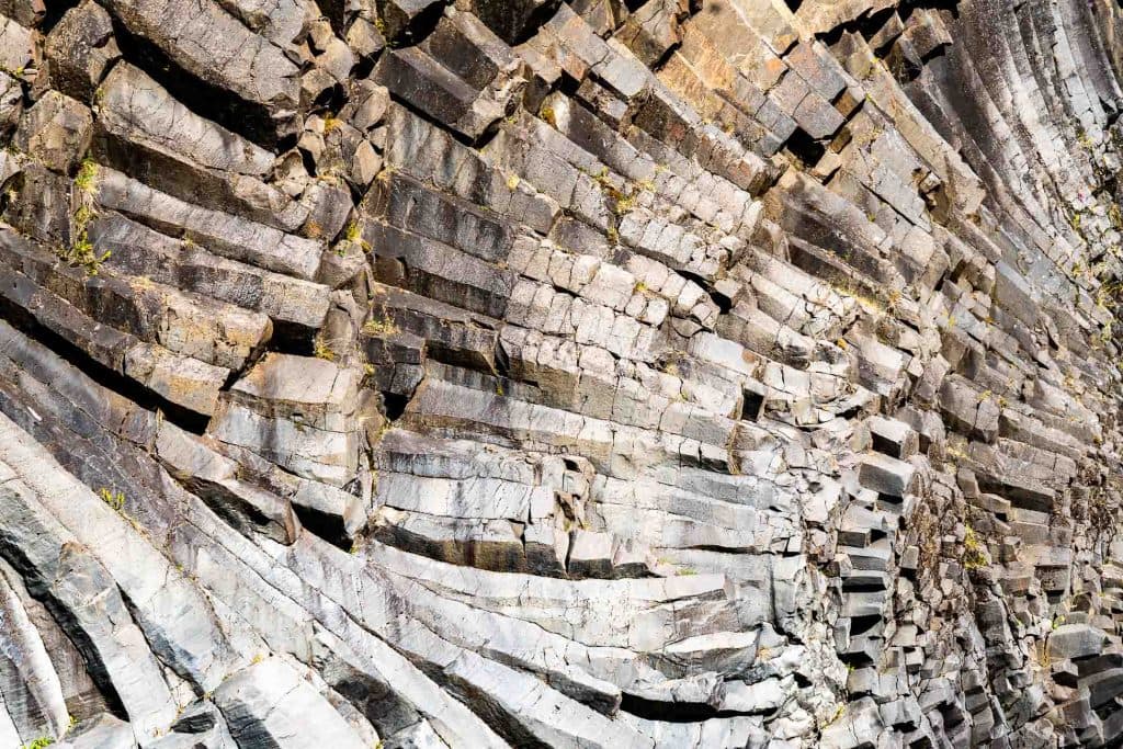 Close-up van een rotsformatie met zeshoekige basaltzuilen in een gebogen, waaiervormig patroon - een iconisch gezicht voor IJslandnomaden op een 7-daagse roadtrip, met grijze tinten en mos in de spleten.