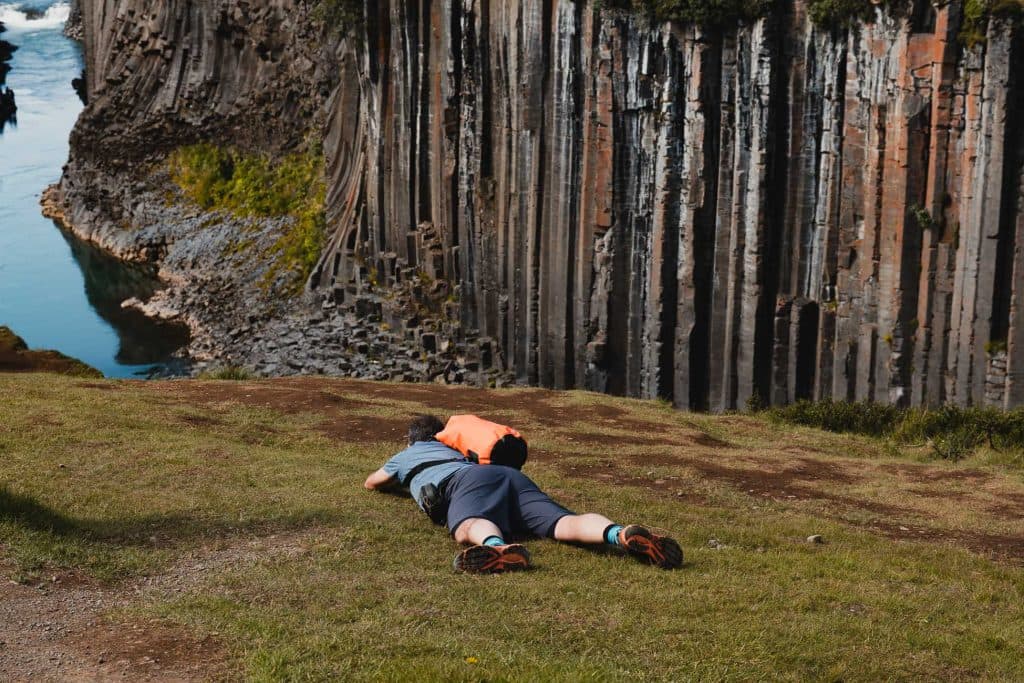 Twee mensen liggen op hun buik op een grasveld en kijken uit over de rand van een klif met dramatische basaltzuilen en een blauwe rivier eronder - een adembenemende stop tijdens een 7-daagse roadtrip met Iceland Nomads.