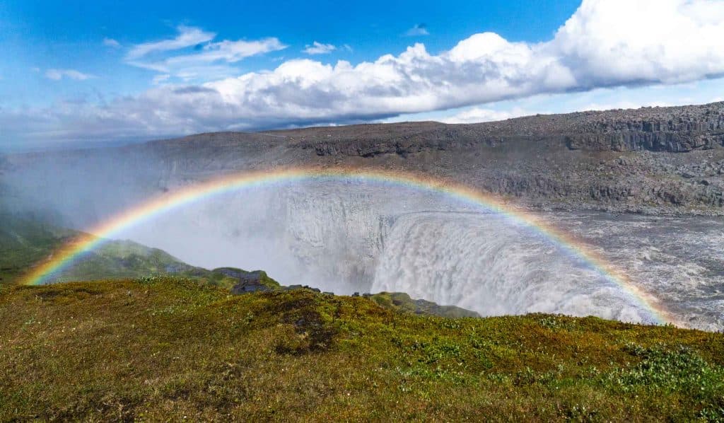 Een levendige regenboog strekt zich uit over een krachtige waterval omringd door rotsachtige kliffen en groen gras, onder een gedeeltelijk bewolkte blauwe hemel - een iconisch gezicht tijdens een 7-daagse Roadtrip IJsland met Iceland Nomads.