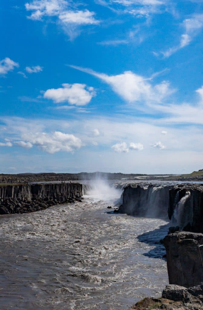Een brede rivier stroomt krachtig over rotsachtige kliffen en vormt watervallen onder een helderblauwe hemel met verspreide wolken - een iconisch tafereel voor elke 7-daagse roadtrip met Iceland Nomads. Mist stijgt op als het water stroomt, omlijst door ruige, rotsachtige oevers.