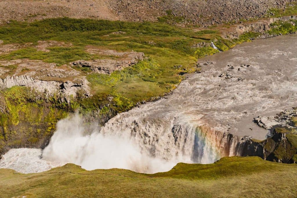 Een grote, krachtige waterval omringd door groene bemoste kliffen en rotsachtig terrein, met oprijzende mist en een zwakke regenboog zichtbaar in de nevel - een must-see hoogtepunt op elke Roadtrip IJsland met Iceland Nomads.