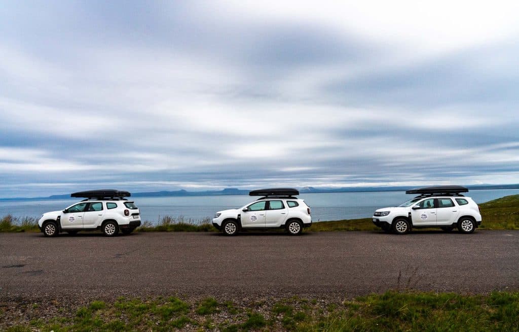 Drie witte SUV's met laadbakken op het dak, die deel uitmaken van een 7-daagse Roadtrip IJsland van Iceland Nomads, staan naast elkaar geparkeerd langs een weg met uitzicht op een kalme zee onder een bewolkte hemel, met groene heuvels en bergen in de verte op de achtergrond.