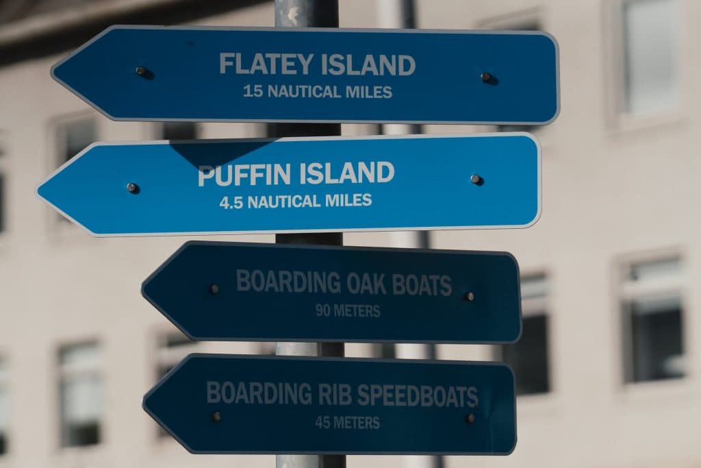 Vier blauwe richtingborden markeren belangrijke plekken voor IJsland Nomaden, waaronder Flatey Eiland (15 zeemijl), Puffin Eiland en lokale bootaanlegplaatsen - perfecte waypoints tijdens je Roadtrip IJsland avontuur. Op de achtergrond staat een gebouw.