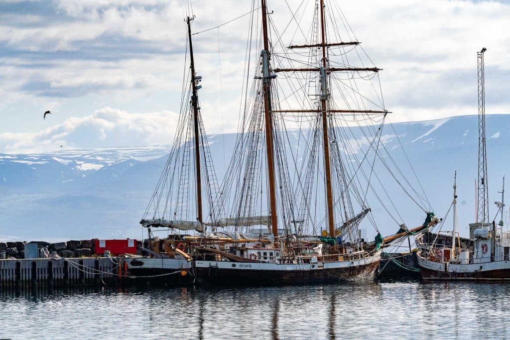 Een grote houten zeilboot met meerdere masten en tuigage ligt aangemeerd in een haven, met kalm water op de voorgrond en besneeuwde bergen erachter - een iconisch tafereel dat perfect is voor elke IJsland Nomads of een 7-daagse roadtrip avontuur.