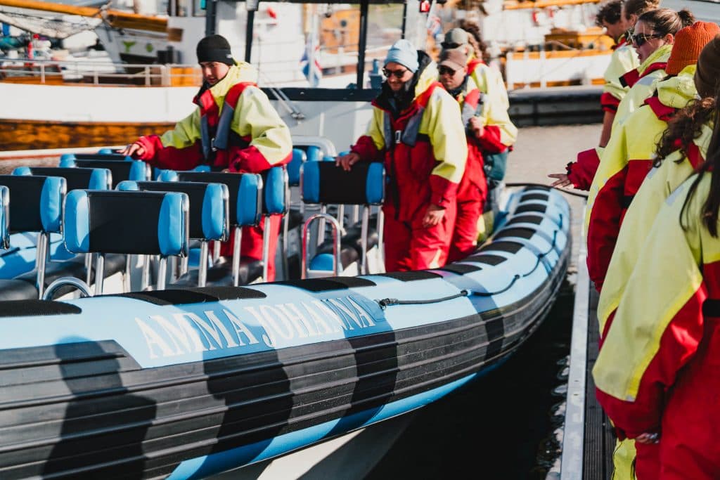 Een groep mensen in rode en gele pakken stapt aan boord van een blauw met zwarte opblaasboot genaamd "Anna Johanna" bij een aanlegsteiger. Dit geeft de avontuurlijke geest weer van een Iceland Nomads 7-daagse roadtrip, met andere boten en mensen op de achtergrond.