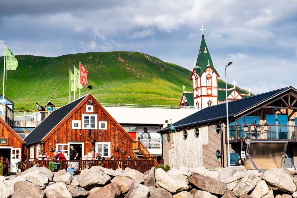 Kleurrijke houten gebouwen en een kerk met een groen dak steken af tegen een met gras begroeide heuvel en een bewolkte hemel, terwijl mensen buiten ontspannen - een uitnodigend tafereel dat typisch is voor een Iceland Nomads 7-daagse roadtrip, met stenen rotsen op de voorgrond.