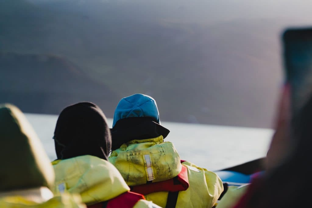Twee mensen met gele jassen en capuchons zitten in een boot en staren naar het water terwijl zacht zonlicht de bergen in de verte baadt - een sereen moment tijdens een 7-daagse roadtrip met Iceland Nomads.