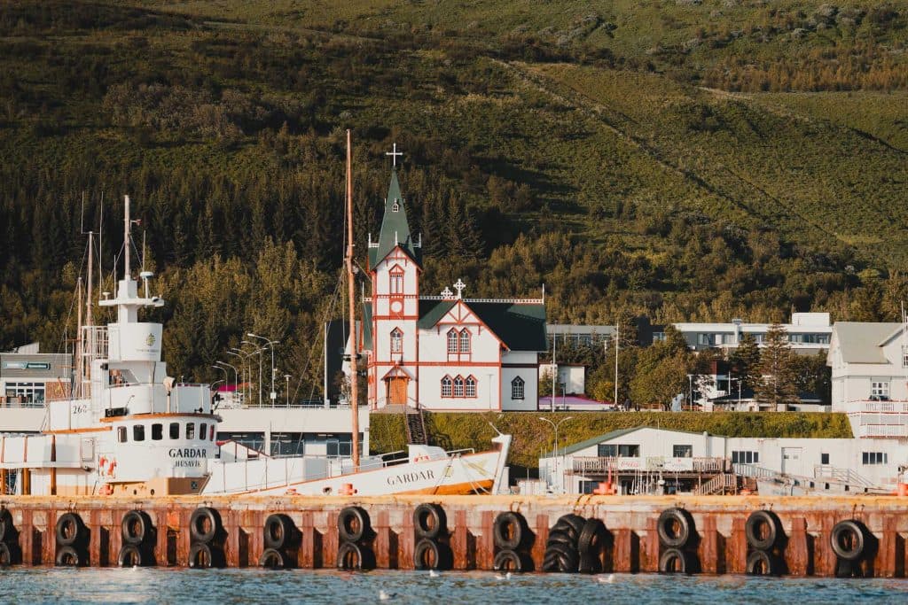 Een witte en groene kerk met een centrale torenspits staat vlak bij het water, omringd door bomen en gebouwen - een idyllisch tafereel voor IJslandse Nomaden op een 7-daagse roadtrip, met vissersboten aangemeerd bij de pier en groene heuvels die oprijzen op de achtergrond.