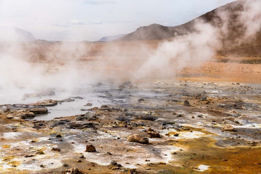 Stoom stijgt op uit borrelende geothermische warmwaterbronnen en verspreide rotsen in een kaal, rotsachtig landschap met bergen in de verte onder een bewolkte hemel - het perfecte decor voor een IJsland Nomads 7-Daagse Roadtrip IJsland avontuur.