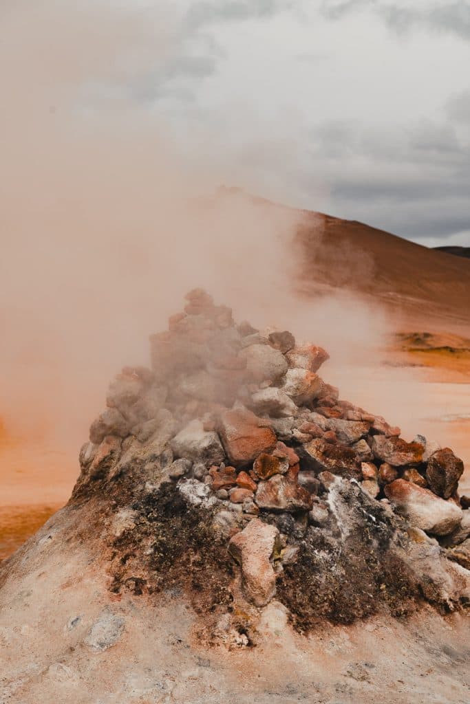 Een dampende stapel rotsen stoot dikke witte damp uit in een dor, rotsachtig landschap - perfect voor een IJsland Nomads avontuur tijdens uw 7-daagse roadtrip onder de uitgestrekte, bewolkte hemel.