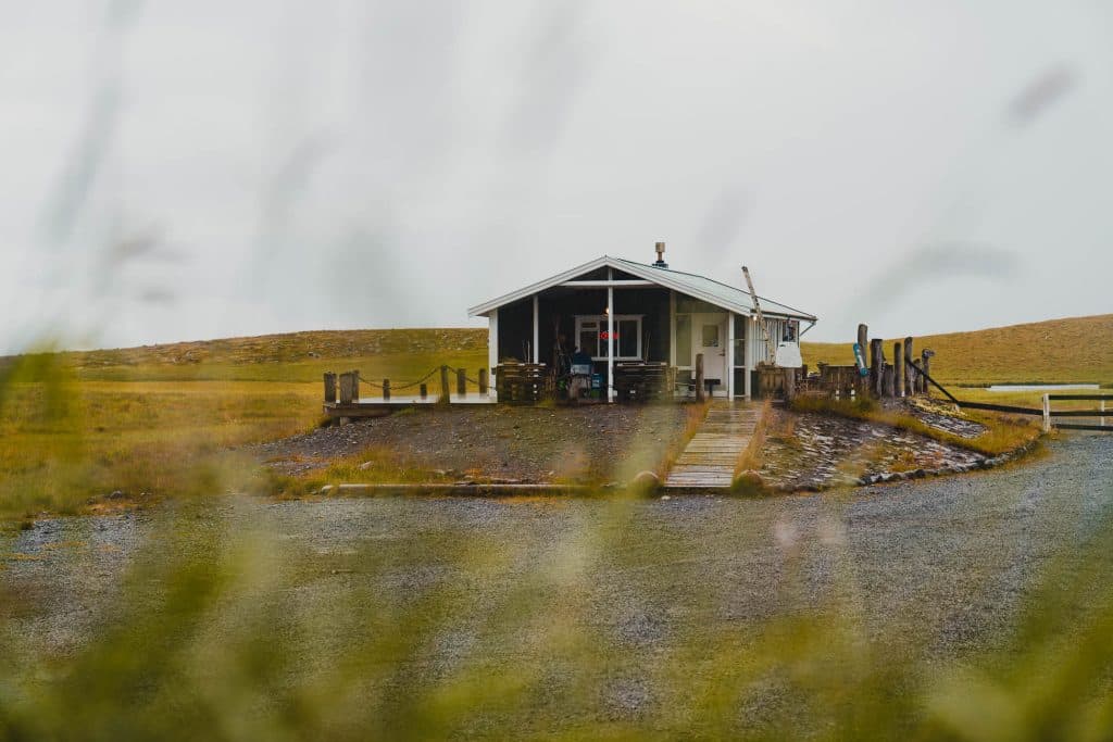 Een klein wit hutje met een veranda staat op een open graslandschap onder een bewolkte hemel, met hoge grassprieten onscherp op de voorgrond - de perfecte scène voor uw 7-daagse roadtrip met Iceland Nomads.