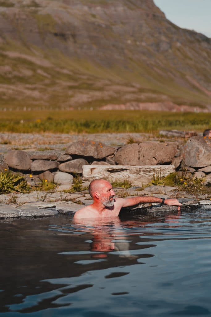 Een kale, bebaarde man ontspant in een warmwaterbron omringd door rotsen en gras, met bergen op de achtergrond onder een heldere hemel - een idyllisch moment voor IJslandse Nomaden op een 7-daagse roadtrip door adembenemende landschappen.