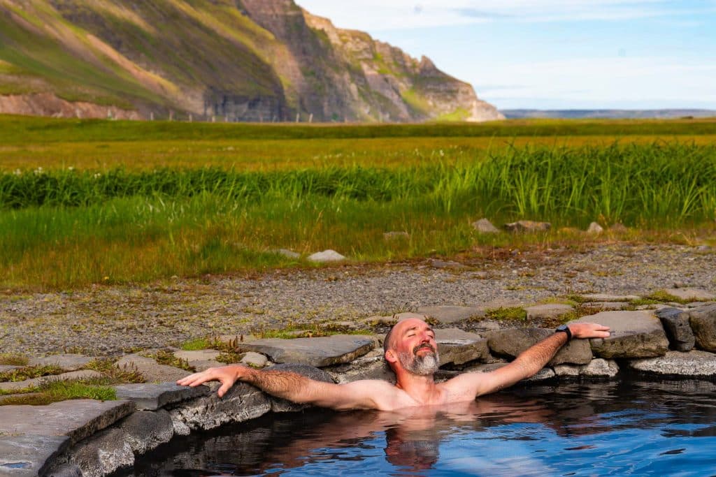 Een man ontspant met uitgestrekte armen in een warmwaterbron, omringd door groen gras en rotsachtige bergen onder een blauwe hemel - perfect voor uw Iceland Nomads 7-daagse roadtrip avontuur.