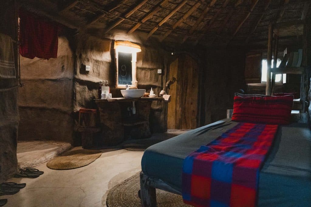 Een rustieke kamer in Afrika met aarden muren en een rieten dak, met een bed met een rood en blauw geruite deken, een houten paaldeur Tanzania, plus een badmeubel met ronde wastafel, spiegel en warme verlichting.