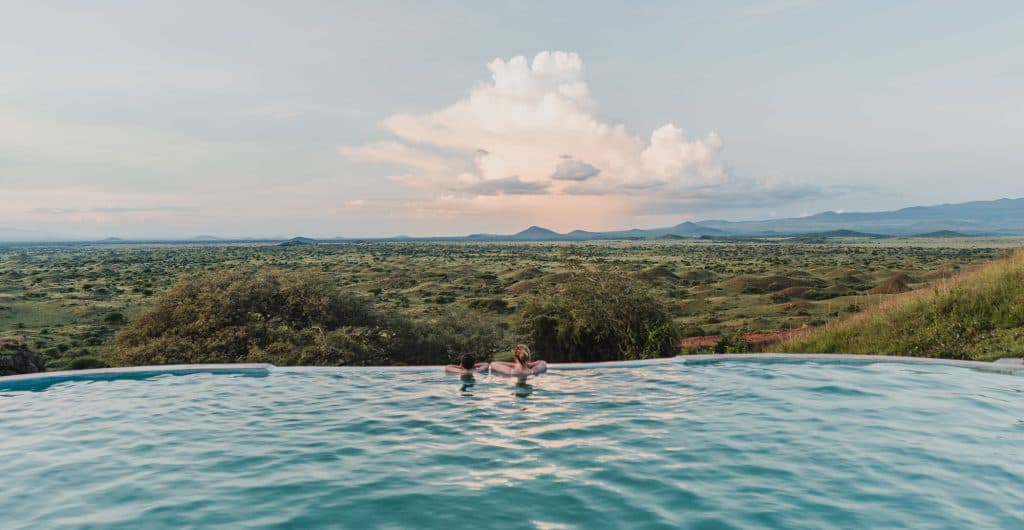 Twee mensen ontspannen in een overloopzwembad met uitzicht op een uitgestrekte groene savanne in Afrika, verspreide bomen en bergen in de verte onder een dramatische zonsondergang hemelpool door Tanzania.
