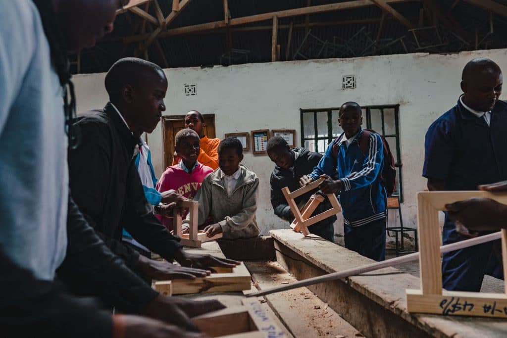 Een groep mensen in een werkplaats schuurt en monteert houten frames voor het Pole pole door Tanzania project, verzameld rond een werkbank in een klaslokaalachtige omgeving met gereedschap en houten balken zichtbaar.