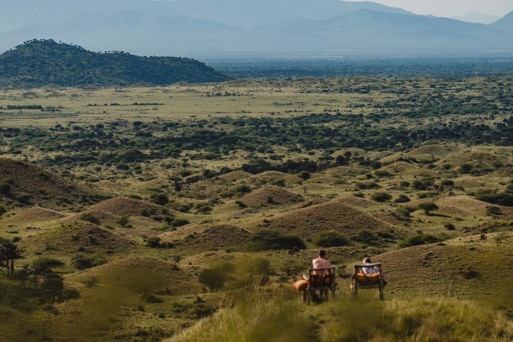 Twee mensen zitten in een stoel op een met gras begroeide heuvel in Afrika, met uitzicht op een uitgestrekt, glooiend landschap bezaaid met struiken en bergen in de verte onder een wazige hemel - een uitnodigend tafereel dat doet denken aan pooldeur Tanzania.