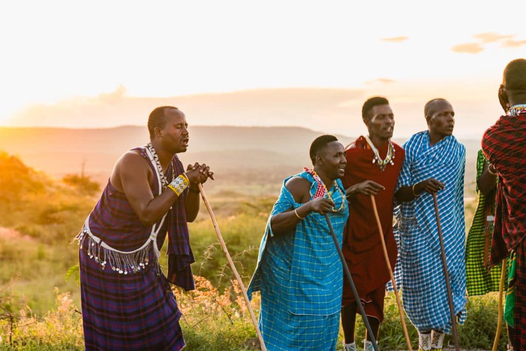 Vijf Maasai mannen in traditionele kleurrijke gewaden en kralenwerk staan samen buiten bij zonsondergang in Tanzania, met houten staven in de hand, omringd door grasvlakten en heuvels in Afrika in de verte.
