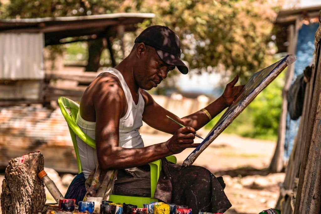 Een man in een witte tanktop en een zwarte pet zit buiten op een groene plastic stoel in Afrika op een doek te schilderen. Gefocust op zijn werk te midden van verfpotten, bomen en rustieke structuren, vangt hij de geest van de artistieke scène van Tanzania.